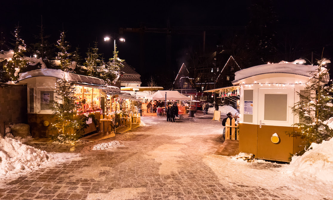 Mercatino di Natale illuminato di sera, con casette in legno e neve sul Renon