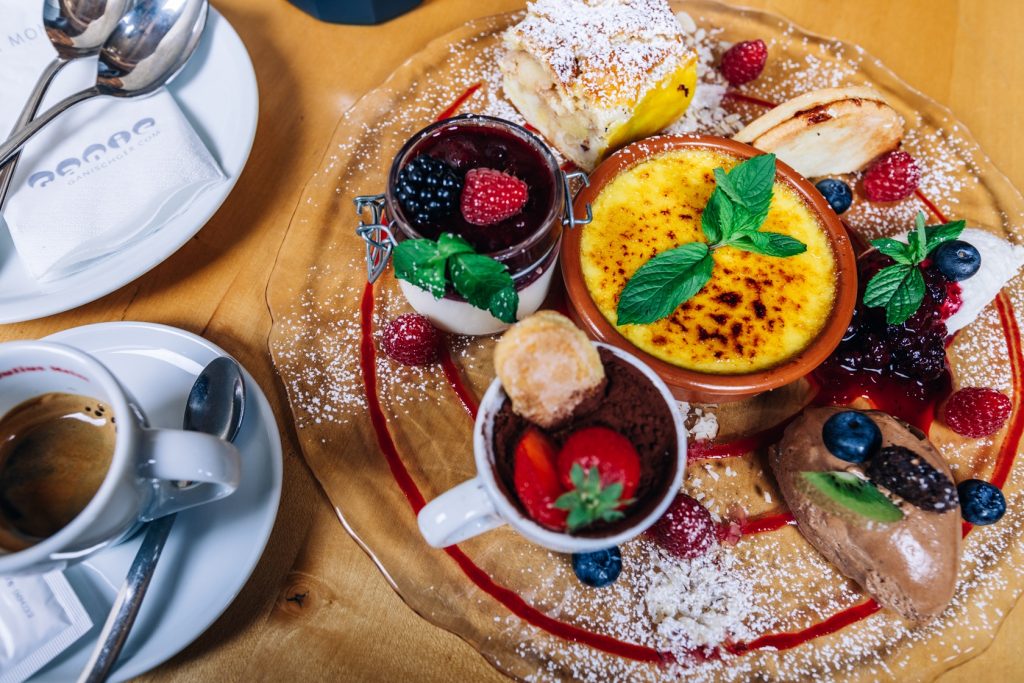 Piatto di dolci con frutti di bosco, creme e gelato, accanto a una tazza di caffè.