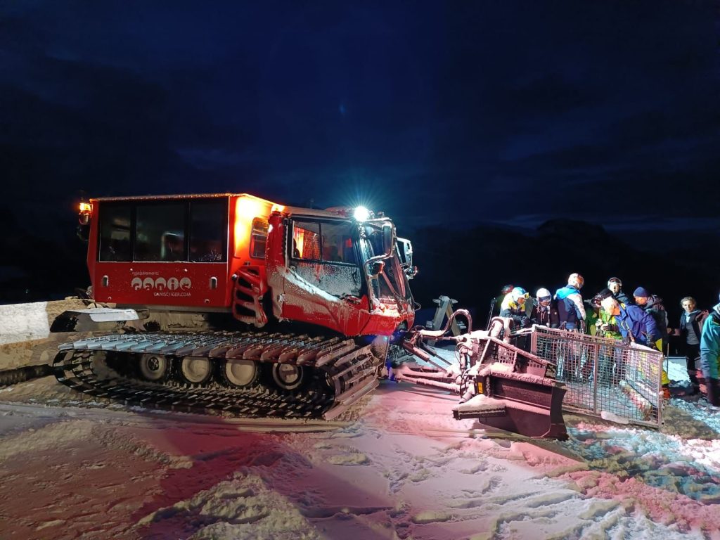 Gatto delle nevi rosso illuminato al buio, con un gruppo di persone accanto.