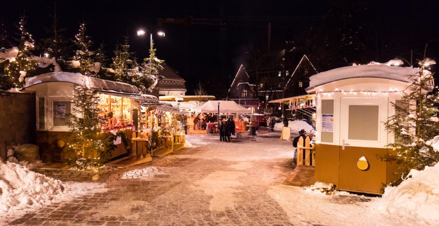 Mercatino di Natale illuminato di sera, con casette in legno e neve sul Renon